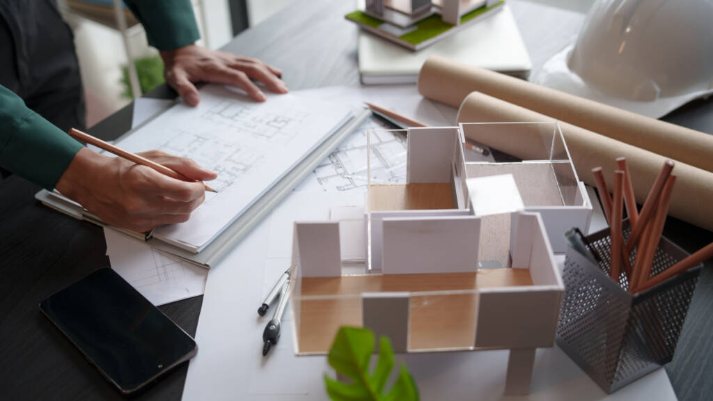 Architect working on building plan with model and blueprint on office desk.